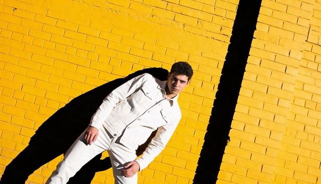 Sunlit male model leaning on yellow brick wall in white denim with dramatic shadow contrast