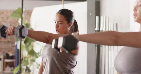 Women performing front raises with hex dumbbells in industrial gym studio for shoulder strength