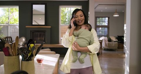 Mother speaking on smartphone while holding baby in cozy kitchen