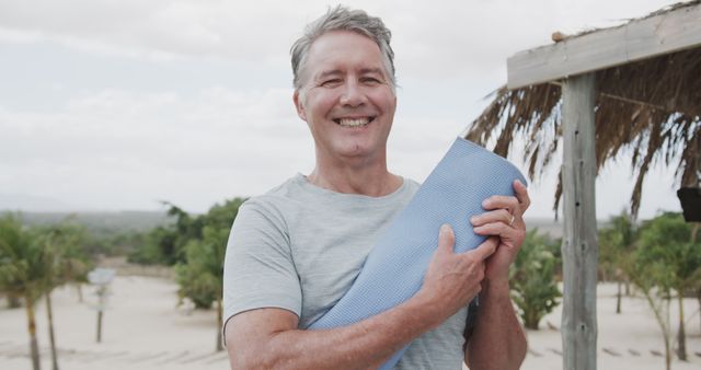 Middle-aged man smiling and holding a yoga mat outdoors at a beach setting. Can be used for promoting wellness, fitness programs, active lifestyle and healthy living. Suitable for advertisements, health blogs, and social media posts related to exercise and outdoor activities.