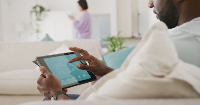 Man comfortably using tablet to manage smart home devices from couch, living room background. Perfect imagery for tech articles, home automation guides, smart living features, and interior design insights.