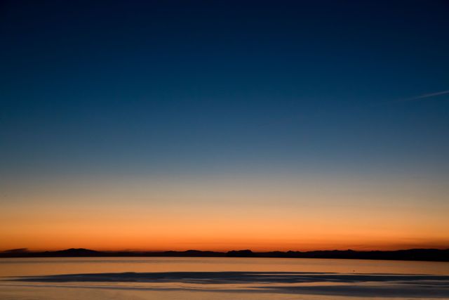 Serene sunset scene with a calm sea reflecting the vibrant colors of the setting sun. Mountains are visible in distance under clear sky. Perfect for backgrounds, travel blogs, wellness content, and meditation visuals.