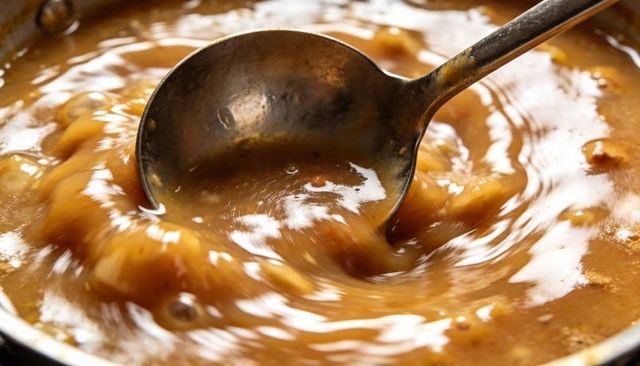 Stirring rustic brown gravy with metal ladle creating glossy swirls and bubbles closeup