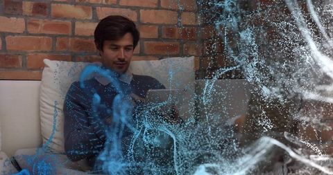 Mid-adult man sitting on sofa using laptop with blue particle overlay, brick lounge
