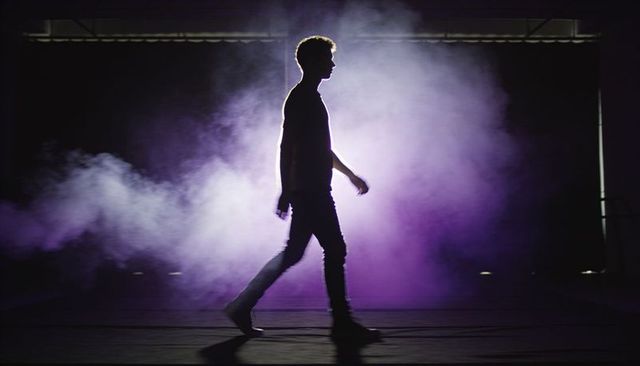 Silhouetted performer walking across smoke-filled stage with purple and white backlight