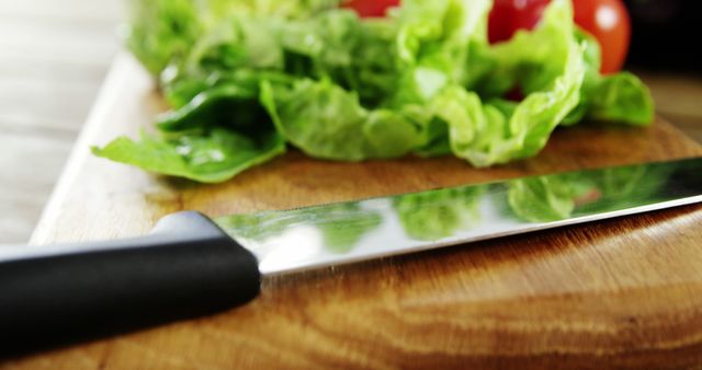 A sharp kitchen knife rests on a wooden cutting board next to fresh lettuce and tomatoes, with copy space. The scene suggests the preparation of a healthy meal, a salad.