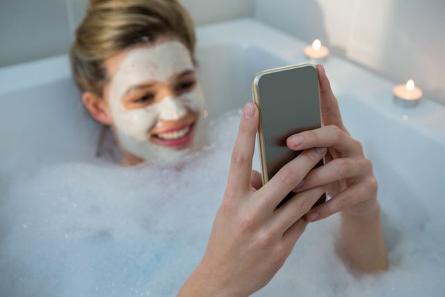 Woman enjoying a relaxing bubble bath while using a mobile phone. She has a face mask on and is surrounded by lit candles, creating a spa-like atmosphere. Ideal for concepts related to self-care, relaxation, technology use, and home spa treatments.