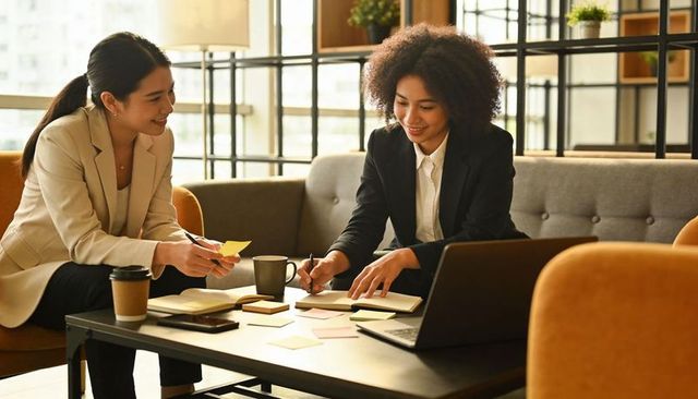 Two women collaborating in modern office lounge writing sticky notes and planning strategy