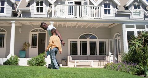 African american father and son playing soccer on front lawn of modern suburban home