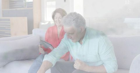 Mature couple relaxing on sunlit modern sofa using tablet and reaching toward coffee table