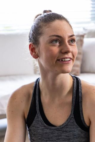 Young woman gazing upward in sunlit living room wearing workout tank top and casual activewear