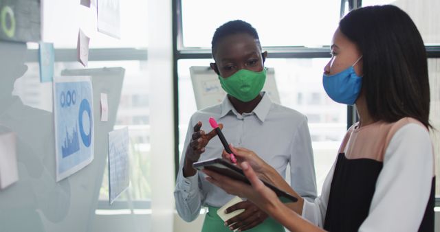 Diverse businesswomen discussing charts on whiteboard while wearing face masks in office - Download Free Stock Images Pikwizard.com