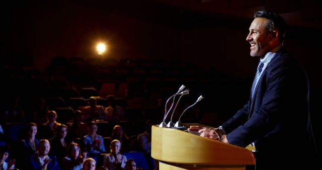 Business professional confidently addressing audience from a lectern in a dimly lit auditorium. Suitable for use in articles on public speaking, leadership training, business seminars, and presentation skills.