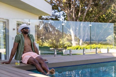 African american man relaxing poolside on modern deck wearing hat and sunglasses with drink