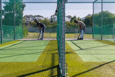 Two cricketers practicing batting in adjacent outdoor nets on sunny training ground