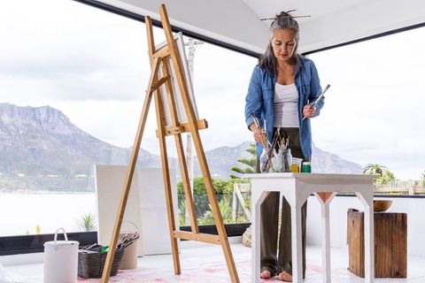 Mature woman painting in minimalist lakeside studio barefoot mixing paints by easel