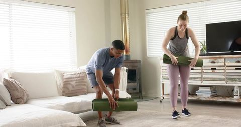 Diverse couple placing green yoga mats in bright living room prepping for home workout