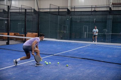 Indoor padel players practicing on blue court collecting balls near net with glass backdrop