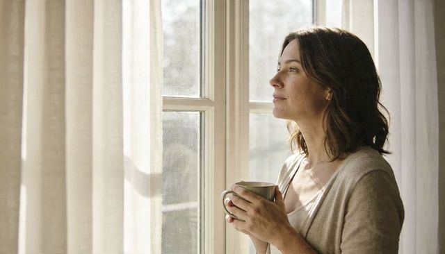 Morning coffee moment woman holding mug gazing through sunlit window in cozy neutral home