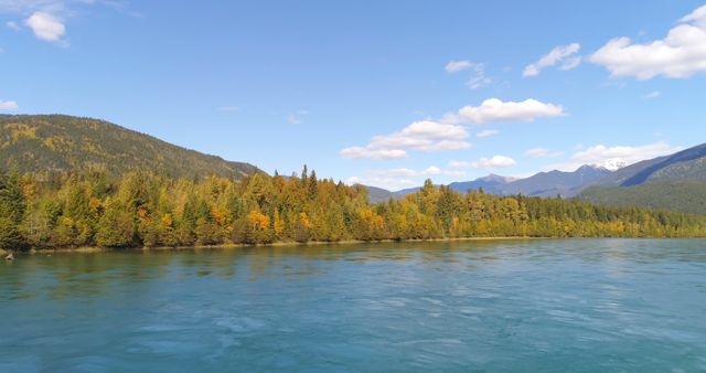 This shows a beautiful autumn scene with a calm river surrounded by a forest showing fall foliage, and a mountain range beneath a clear blue sky. Perfect for promoting travel destinations, outdoor activities, nature retreats, and seasonal backgrounds. Ideal for use in brochures, websites, and social media related to outdoor excursions and environmental beauty.