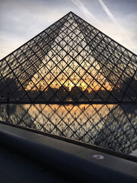 Elegant glass pyramid of Louvre Museum in Paris during sunset, showcasing architectural beauty. Soft twilight sky enhances ambiance with golden reflections on water. Ideal for travel blogs, tourism promotions, architectural editorials, and cultural posts.