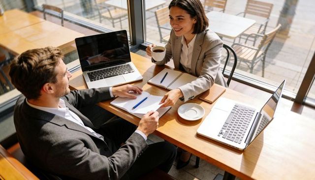 Cafe Meeting: Two Professionals Reviewing Notes and Working on Laptops with Coffee