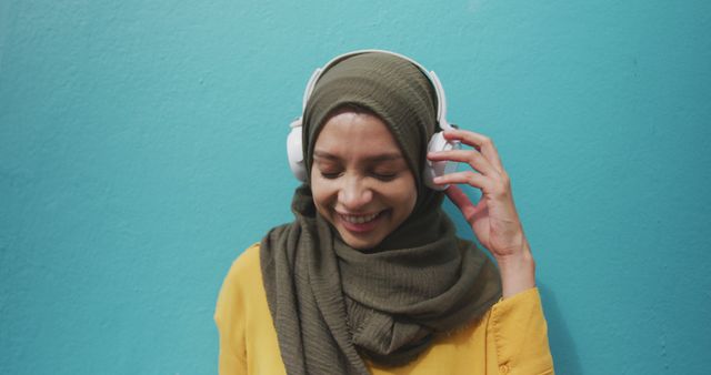 Smiling biracial woman in hijab standing by blue wall listening to music on headphones. City living, music and modern urban lifestyle.