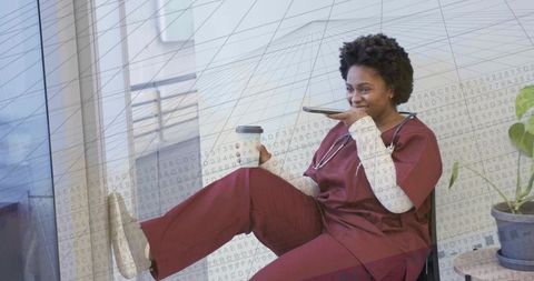 Black nurse taking break with coffee and smartphone in clinic wearing burgundy scrubs