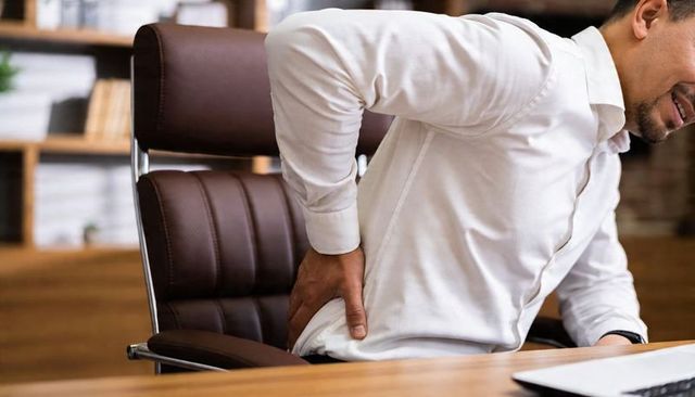 Office worker holding lower back while sitting at desk showing lumbar pain from poor ergonomics