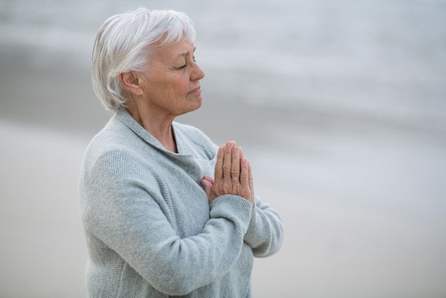 Senior woman standing on beach with hands in prayer position, eyes closed, and serene expression. Ideal for use in articles or advertisements related to spirituality, mindfulness, meditation, relaxation, and senior wellness. Can also be used for promoting peaceful lifestyles and nature retreats.