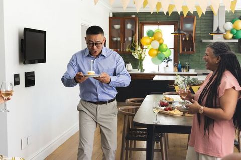 Senior celebrating birthday at home holding cupcake with candle, friends cheering in kitchen