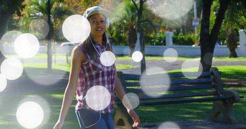 Young woman walking in sunny park wearing headphones and cap, casual bokeh lifestyle
