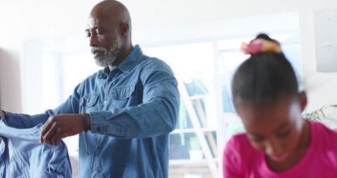 Senior african american father folding shirt while daughter leaning forward in bright home