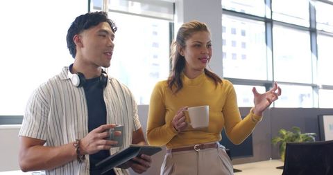 Diverse coworkers discussing project while holding mugs and tablet in modern open office