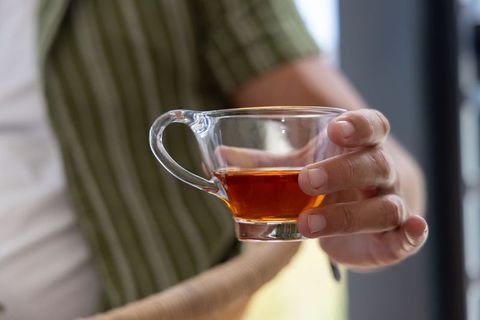 Male holding clear glass teacup with amber tea near bright window, cozy interior