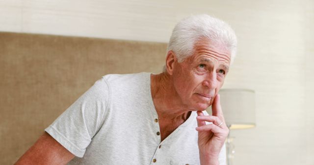 A senior man with white hair appears deep in thought while at home. Wearing a casual gray top, he leans slightly to one side with a hand to his chin, suggesting a moment of contemplation. This image can be used for topics related to aging, retirement, life reflections, or health and wellness of seniors.