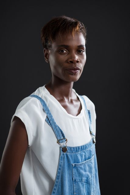 This image features an androgynous individual wearing denim overalls and a white shirt, standing against a grey background. The person exudes confidence and style, making it suitable for use in fashion editorials, gender-neutral clothing advertisements, and modern lifestyle blogs. The neutral background and casual attire highlight the subject's unique expression and contemporary fashion sense.
