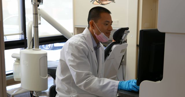 Dentist is focused on computer screen while wearing white lab coat and safety gloves in a modern dental clinic. There is a dental chair and equipment in the background. This can be used to illustrate healthcare technology, professional dental services, patient records management, or environments in medical practices.