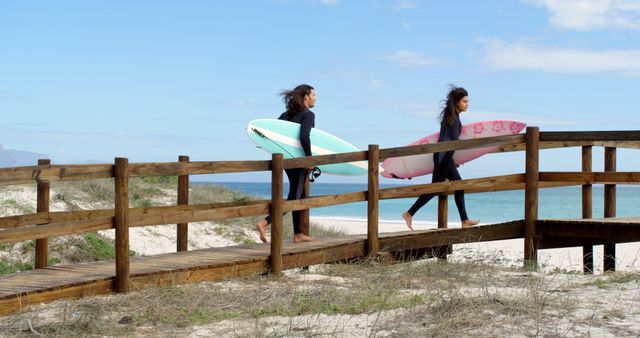Two women carrying surfboards walk on a wooden pathway leading to a sandy beach. They are dressed in wetsuits and are about to enjoy a surfing session in the ocean. Ideal for use in projects related to summer activities, beach vacations, surfing sports, and adventurous lifestyles.