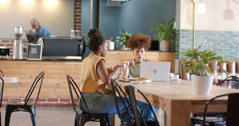 Two women collaborating and discussing over laptop in rustic cafe sharing coffee and pastry