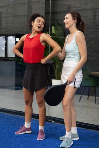 Diverse women chatting on padel court holding paddles in sporty outfits, casual friendship