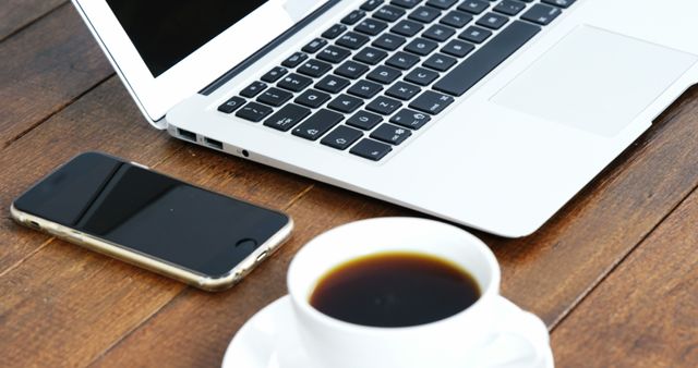 Open laptop and smartphone next to cup of black coffee on sturdy wooden table, embodying a productive modern office setup. Perfect for illustrating remote working, productivity, technology use, or business communication. Ideal for blog posts, websites, or marketing materials focusing on working environments and efficiency tools.