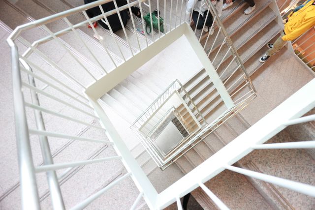 Stunning top view of a modern spiral staircase in a brightly lit building interior. The image features the geometric design and clean lines of the staircase, creating a captivating perspective. Perfect for use in architectural design publications, urban lifestyle blogs, interior design websites, and promotional materials for modern real estate properties.