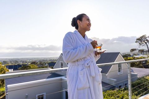 Asian woman in white robe enjoying calm morning tea on coastal balcony overlooking sunrise