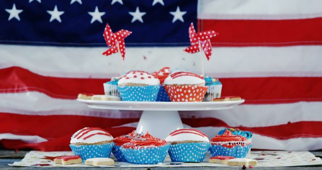 Cupcakes decorated in red, white, and blue are placed on a white stand with an American flag in the background. Red polka-dotted pinwheels add a festive touch. Ideal for use in content about American holidays, festive baking, party planning, or Fourth of July celebrations.