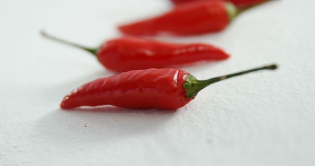 Three red chili peppers are aligned diagonally on a white textured surface, with the focus on the foremost chili. Their vibrant color and sleek shape suggest a spicy addition to culinary dishes.