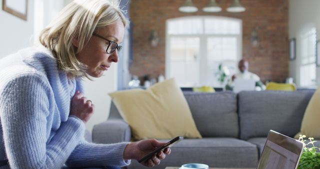 Worried senior caucasian woman in living room sitting on sofa, using smartphone and laptop. retirement lifestyle, at home with technology.