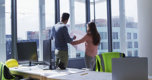 This image shows two architects engaged in a discussion centered around a set of blueprints within a contemporary office space. The large windows reveal a cityscape and bring abundant natural light, enhancing productivity. The desks are equipped with dual monitors, a laptop, desk lamp and safety gear, indicative of a professional and collaborative work environment. This image is ideal for representing themes of teamwork, modern workplaces, engineering projects, and urban planning.