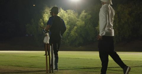 Night cricket batter at crease under floodlights with umpire walking on grass field