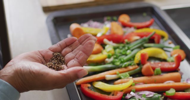 Hand holding peppercorns and seasoning colorful vegetables on a baking tray. Ideal for healthy eating recipes, food blogs, and content about cooking at home. Useful for promoting healthy lifestyle and nutritious meal preparation.